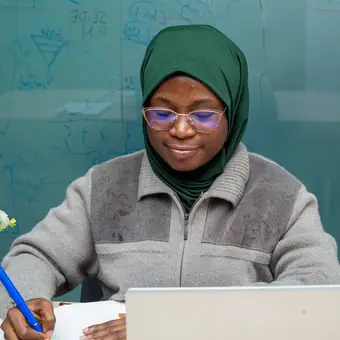 Une femme portant un foulard vert et des lunettes écrit dans un carnet, assise à un bureau