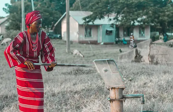 Une femme en tenue traditionnelle rouge actionne une pompe à eau manuelle, un seau se remplit