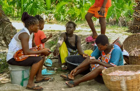 Trois femmes assises à l'ombre discutent près de grands paniers tressés, une enfant à leurs côtés